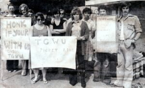 Black-and-white photograph of Ronson workers standing on a pavement during a strike, holding hand-painted TGWU picket line placards and facing the camera.