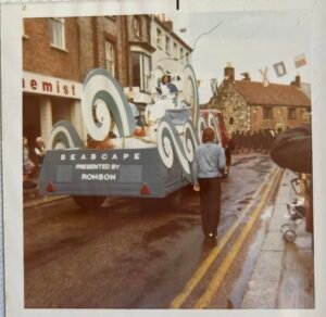 Colour photograph of the rear of the Ronson "Seascape presented by Ronson" carnival float travelling down Newport High Street, with a figure walking alongside and crowds lining the pavement.