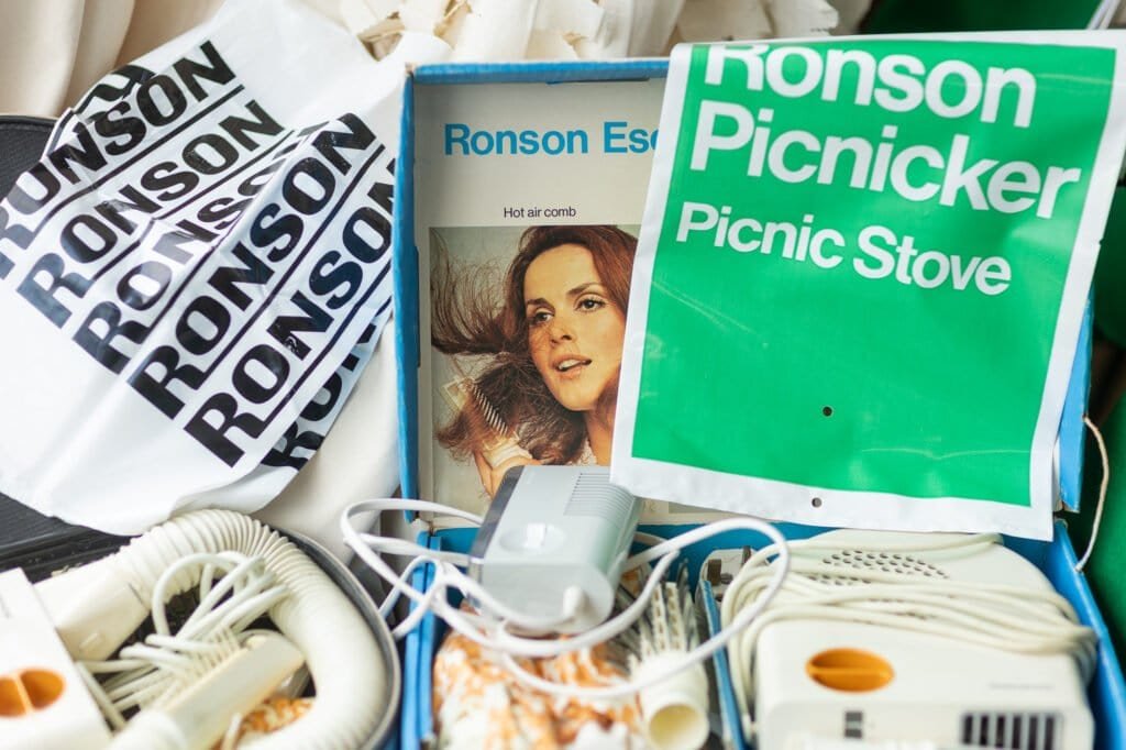 Ronson memorabilia: a Ronson carrier bag, a hot air comb box and a green 'Ronson Picnicker Picnic Stove' sign above white hairdryers.