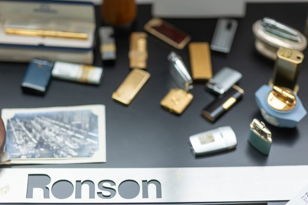 A metal sign with 'Ronson' cut out, in front of a blurred collection of gold, silver and coloured Ronson lighters and an old print.