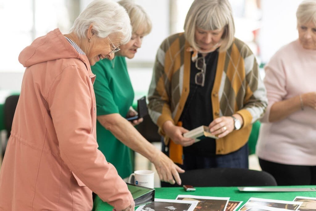 An attendee in a pink jacket laughs and reaches towards printed photos on a table as others look on at the Ronson Reunited event in 2026.