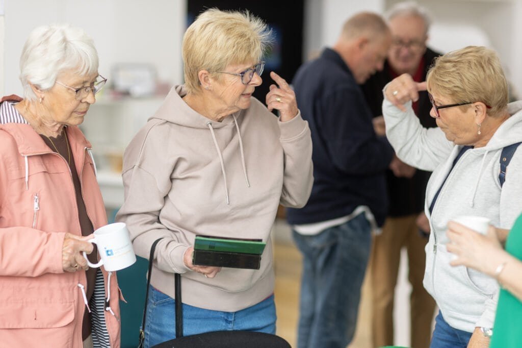 Two attendees look at a small dark green case held by one of them while another gestures towards her head at the Ronson Reunited event in 2026.