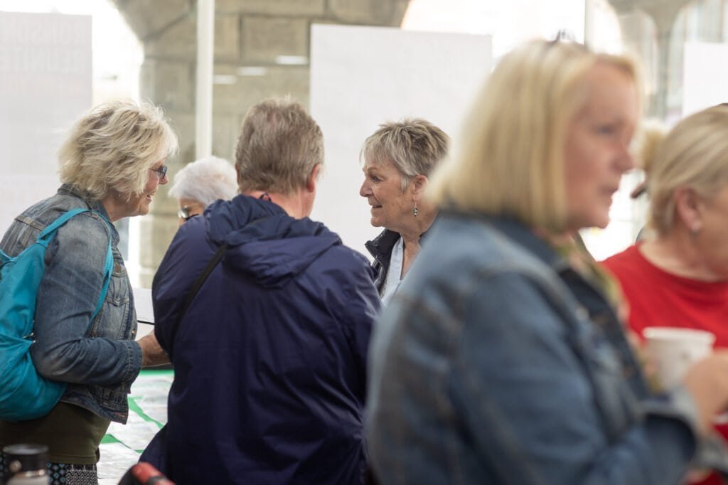 Attendees chat in a group, a smiling woman in a dark gilet talking with a man in a navy jacket seen from behind at the Ronson Reunited event in 2026.