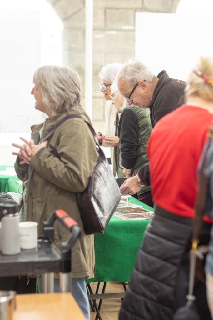 Attendees stand around a table of photos next to a tea station, one in an olive jacket with her hands clasped at the Ronson Reunited event in 2026.