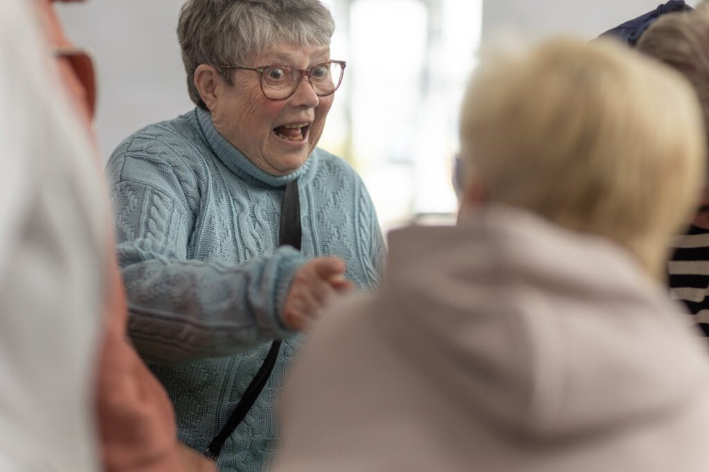 An attendee in a pale blue cable-knit jumper and glasses, mouth open in surprise, reaches out as she speaks at the Ronson Reunited event in 2026.