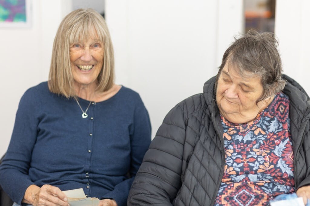 Two attendees sit side by side, one in a navy cardigan smiling broadly and holding small cards at the Ronson Reunited event in 2026.