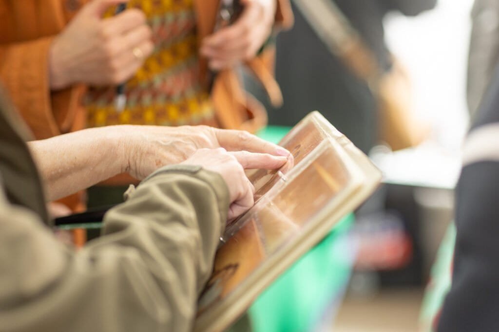 Close-up of hands turning the pages of a photo album with clear plastic sleeves, while others stand behind at the Ronson Reunited event in 2026.