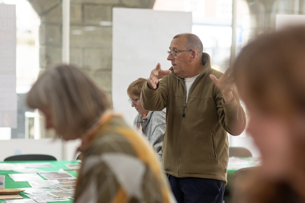 A man in an olive fleece gestures with both hands as he talks, with printed photos on a table behind at the Ronson Reunited event in 2026.