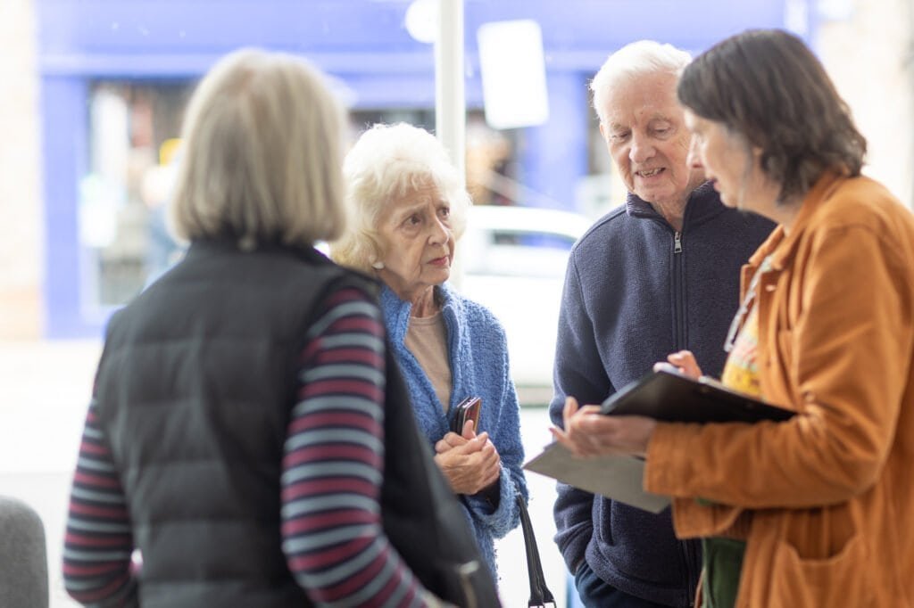 Attendees talk by the entrance, a woman in an orange jacket holding a clipboard speaking with a woman at the Ronson Reunited event in 2026.