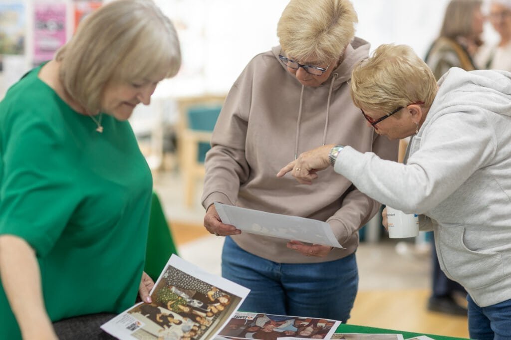 Three attendees lean over a table of printed photos, one holding a printed sheet and another pointing at the Ronson Reunited event in 2026.