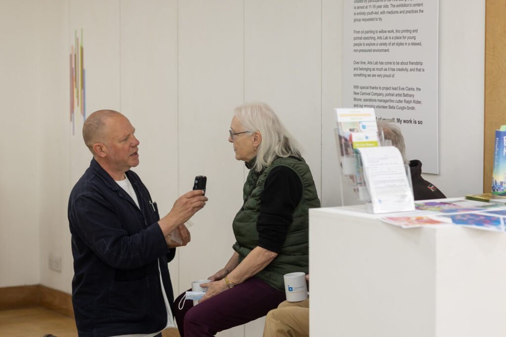 A man holding a handheld recorder as he interviews a seated white-haired woman holding a mug at the Ronson Reunited event in 2026.