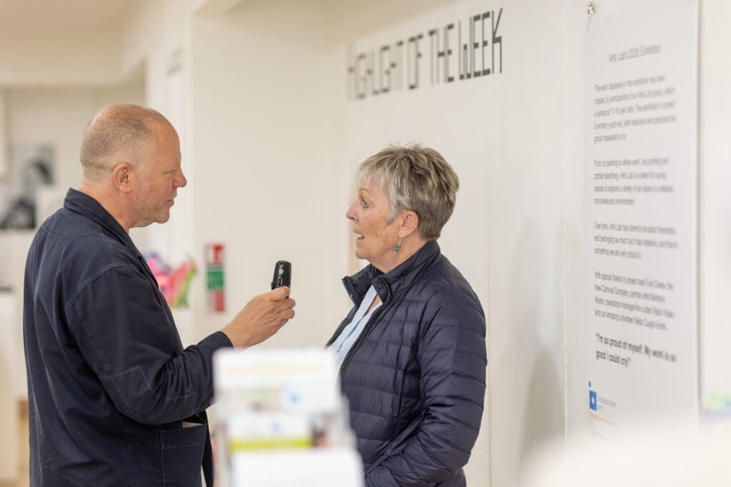 A man holds a handheld recorder towards an attendee in a navy quilted jacket at the Ronson Reunited event in 2026.