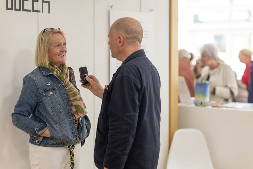A man holds a handheld recorder towards a smiling woman in a denim jacket and scarf at the Ronson Reunited event in 2026.