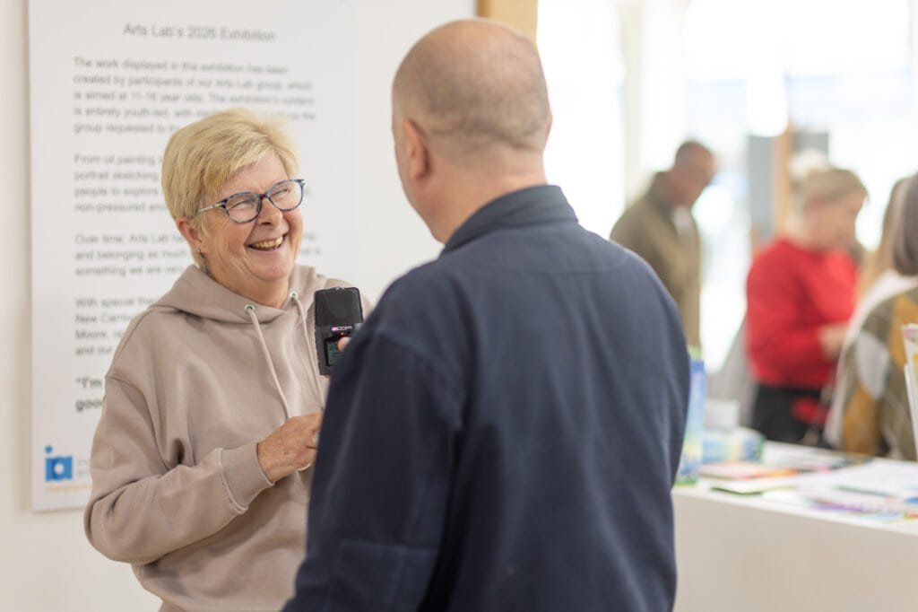 A man holds a handheld recorder towards a laughing woman in glasses and a beige hooded top at the Ronson Reunited event in 2026.