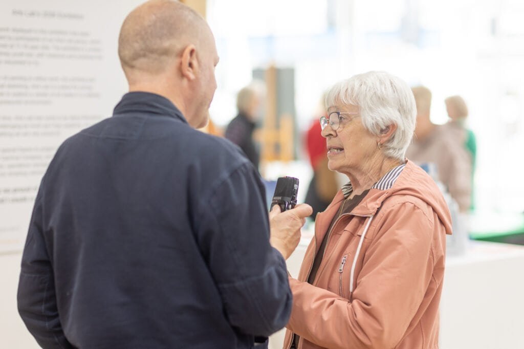 A man holds a handheld recorder towards a woman in glasses and a pink jacket at the Ronson Reunited event in 2026.