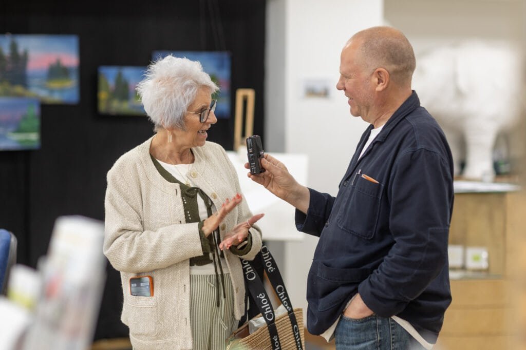 A woman laughing and gesturing with both hands as a smiling man records her on a handheld recorder at the Ronson Reunited event in 2026.